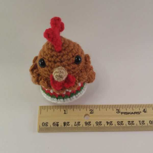 A crochet Christmas chicken behind a ruler with a white background